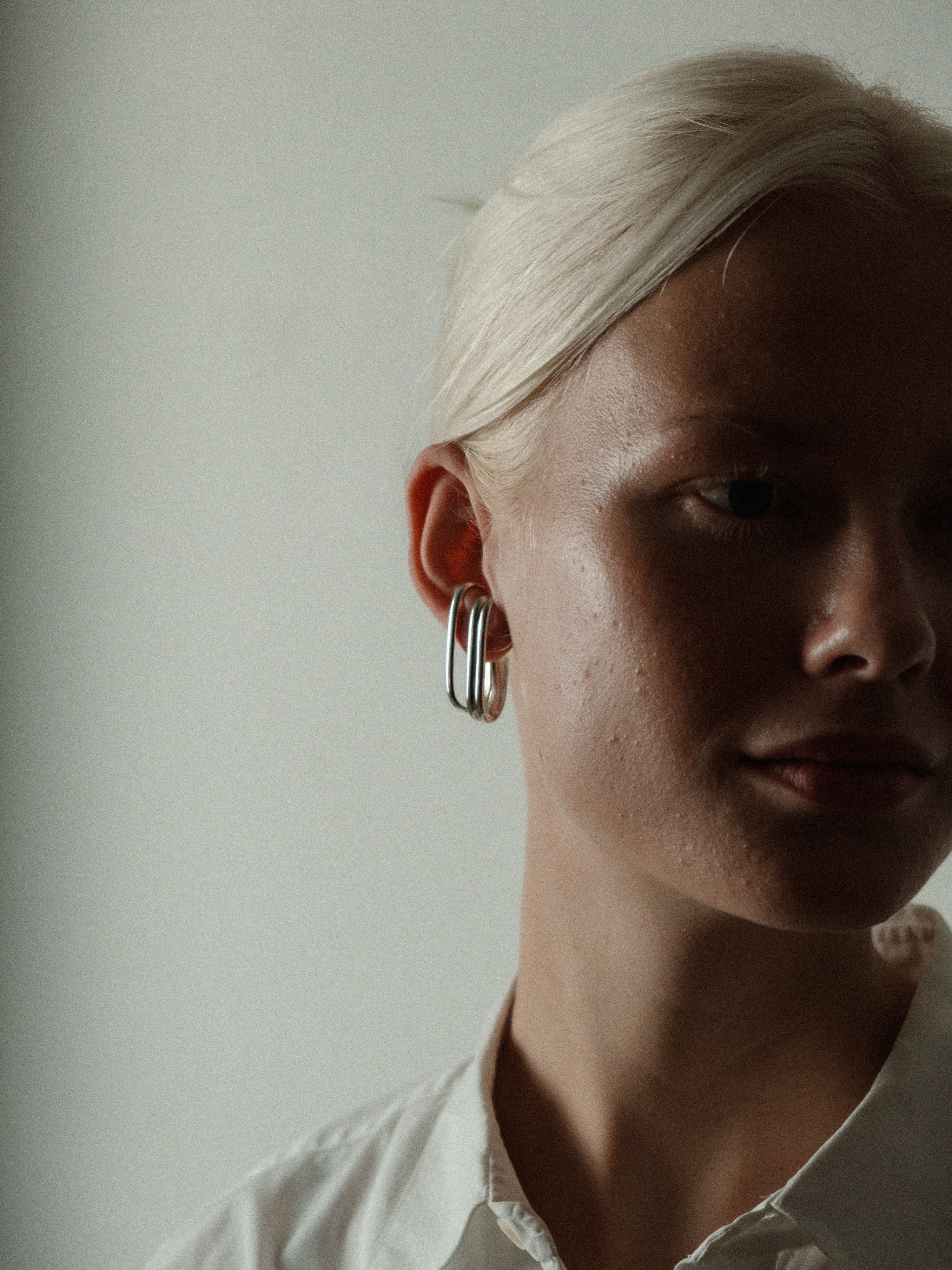 A close-up portrait of a woman with platinum blonde hair wearing a large, silver-toned triple-ring hoop earring, set against a plain, light gray background.
