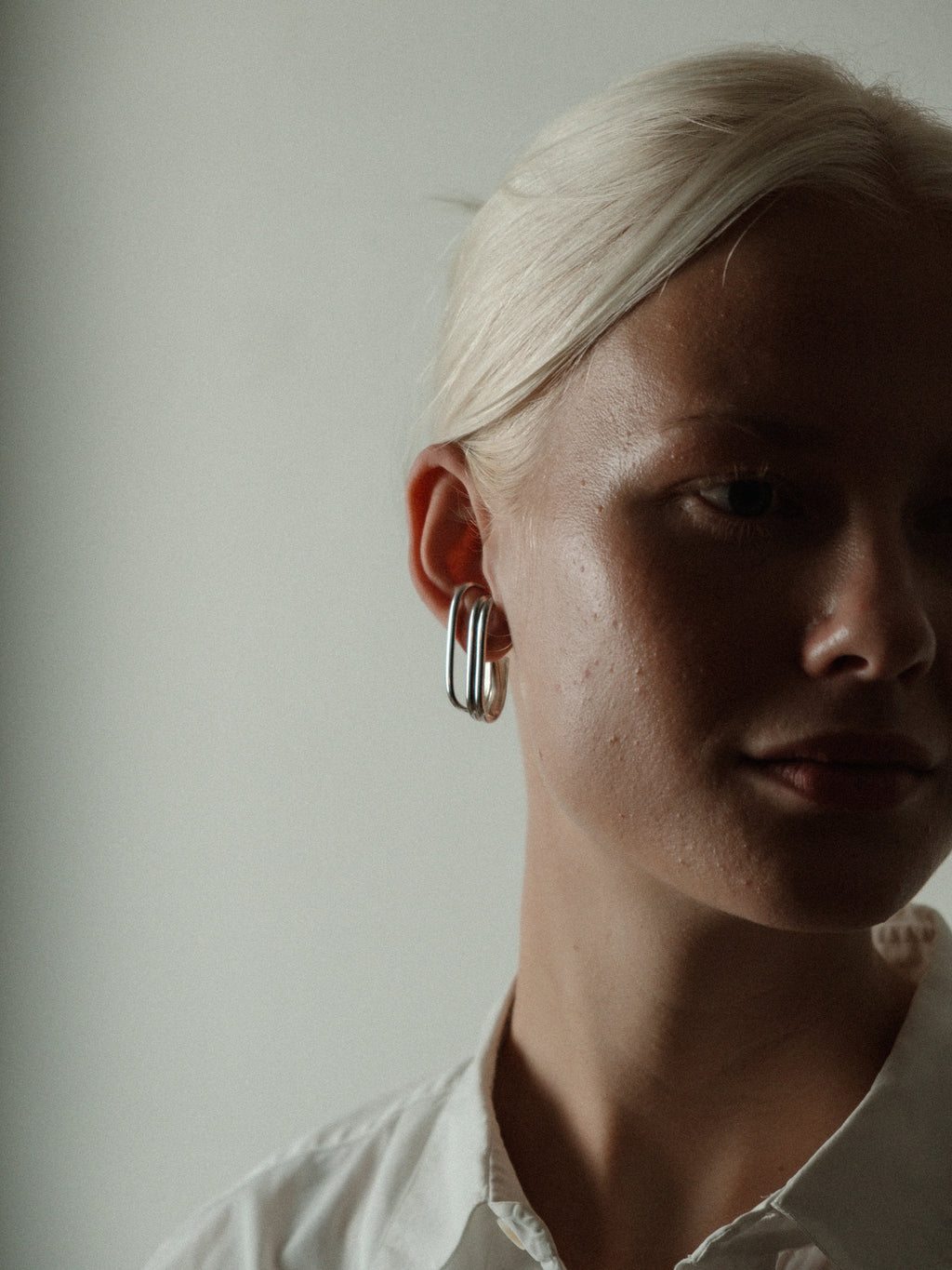 A close-up portrait of a woman with platinum blonde hair wearing a large, silver-toned triple-ring hoop earring, set against a plain, light gray background.