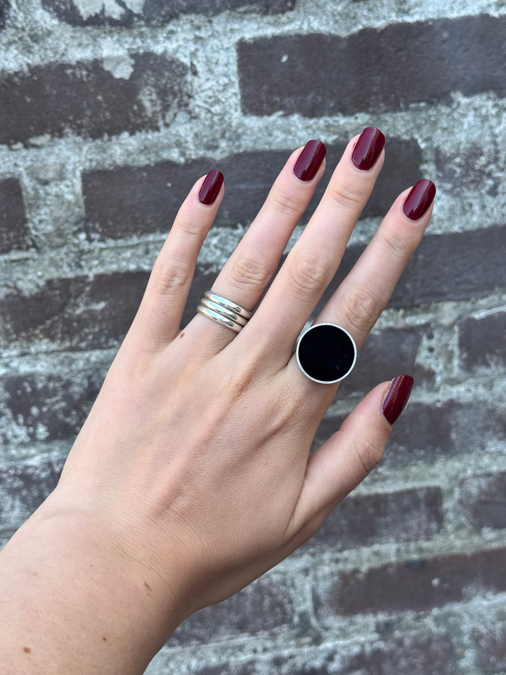 A hand with dark burgundy manicured nails wearing a large silver ring with a black round stone, alongside a thick silver band, set against a gray and white distressed brick background.