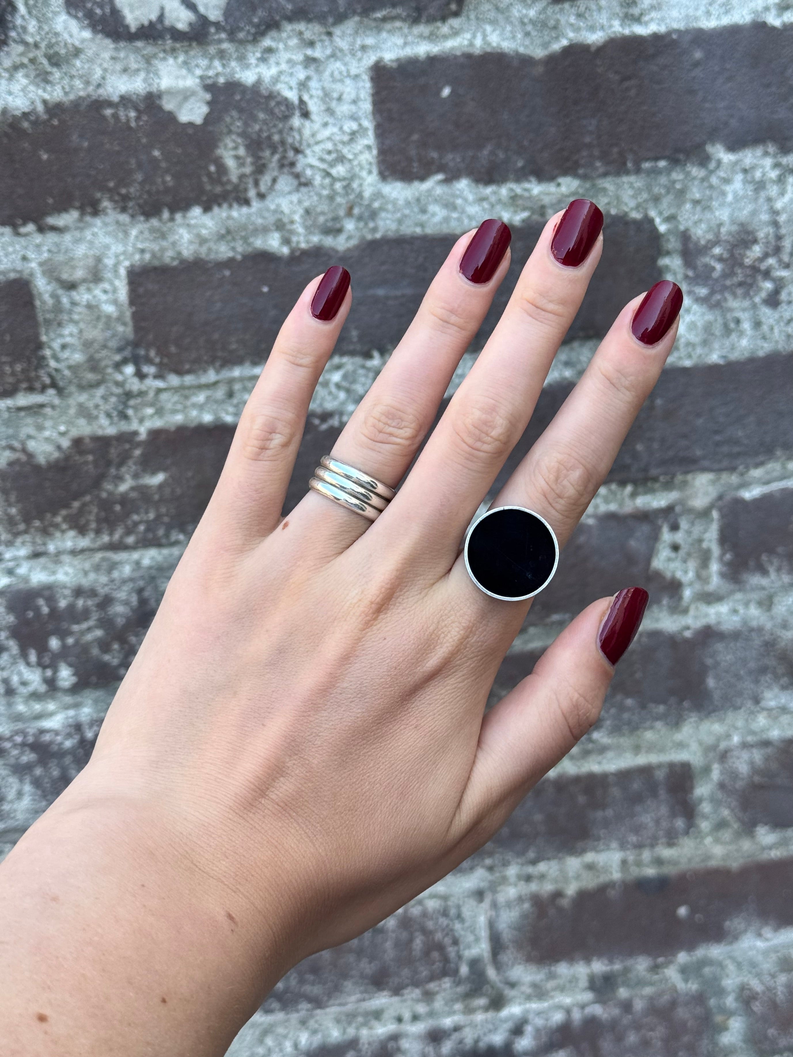 A hand with dark burgundy manicured nails wearing a large silver ring with a black round stone, alongside a thick silver band, set against a gray and white distressed brick background.
