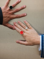 Two hands reaching toward each other against a plain beige background, each wearing a ring. One ring has a black circular face on a silver band, and the other has a bright red circular face on a silver band, with additional silver rings on one hand.